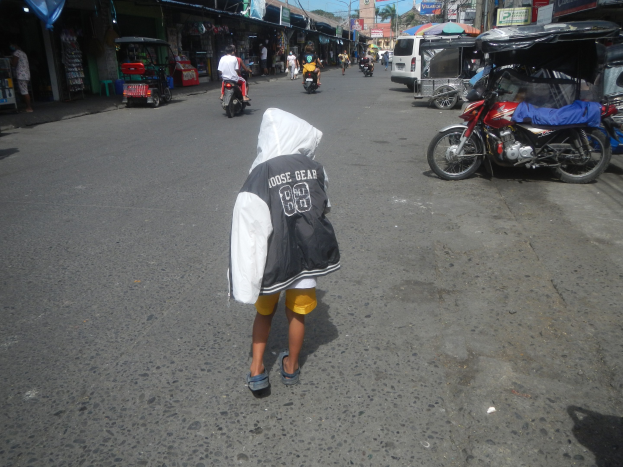 Ein junger Junge geht eine von Motorrädern und anderen Fahrzeugen verstopfte Straße entlang, mit Gebäuden, Namensschildern, Bäumen, Pfählen und einem klaren blauen Himmel im Hintergrund.