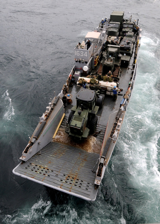 Luftaufnahme eines großen Militärschiffs auf dem Meer mit einem Fahrzeug an Bord, umgeben von Menschen und Gegenständen, mit einem Wasserzeichen unten.
