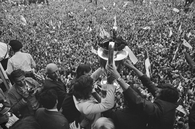 Schwarzes und weißes Foto einer Gruppe von Menschen, die einen Pokal in die Höhe halten, während einige Kameras und Fahnen halten, vor einer großen Menge.