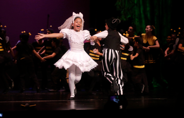 Ein junges Mädchen in einem weißen Kleid und ein Mann in einem schwarzen und weißen gestreiften Anzug tanzen auf einer Bühne, umgeben von kostümierten Menschen, mit einem grünen Vorhang und einer violetten Wand im Hintergrund.