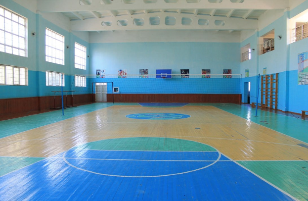 Große Sporthalle mit blau-weißem Boden, Basketballfeld, Netz, Stangen, Tür, Fenstern, Wandplakaten, Geländer und Dach.