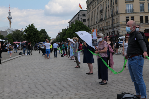Eine Gruppe von Menschen marschiert auf einer Straße in Berlin, Deutschland, mit Schildern und Transparenten, einige tragen Masken und tragen Taschen oder Regenschirme, mit Bäumen, Gebäuden, geparkten Fahrzeugen und einem Flaggenturm im Hintergrund bei bewölktem Himmel.