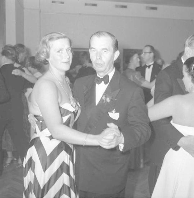 Zwei Menschen tanzen gemeinsam auf einer formellen Veranstaltung, der Mann trägt einen schwarzen Blazer, ein weißes Hemd und schwarze Hosen, hält die Hand seiner Partnerin.