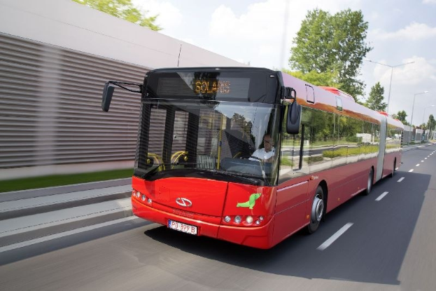 Roter Bus fährt auf einer Straße neben einem Gebäude, mit einer Person im Inneren, neben einer Wand, Bäumen, Laternen und einem bewölkten Himmel.
