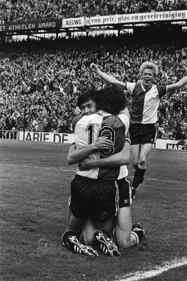 Drei Fussballspieler umarmen sich auf dem Boden beim Feiern eines Tores, mit jubelnder Menge und schwenkenden Fahnen im schwarz-weißen Hintergrund.