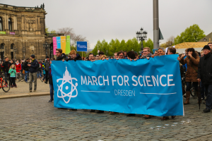 Eine Gruppe von Menschen hält eine "March for Science Dresden"-Fahne vor einem Gebäude mit Fenstern, umgeben von Bäumen, Laternenmasten und Fahrrädern, mit dem Himmel im Hintergrund.