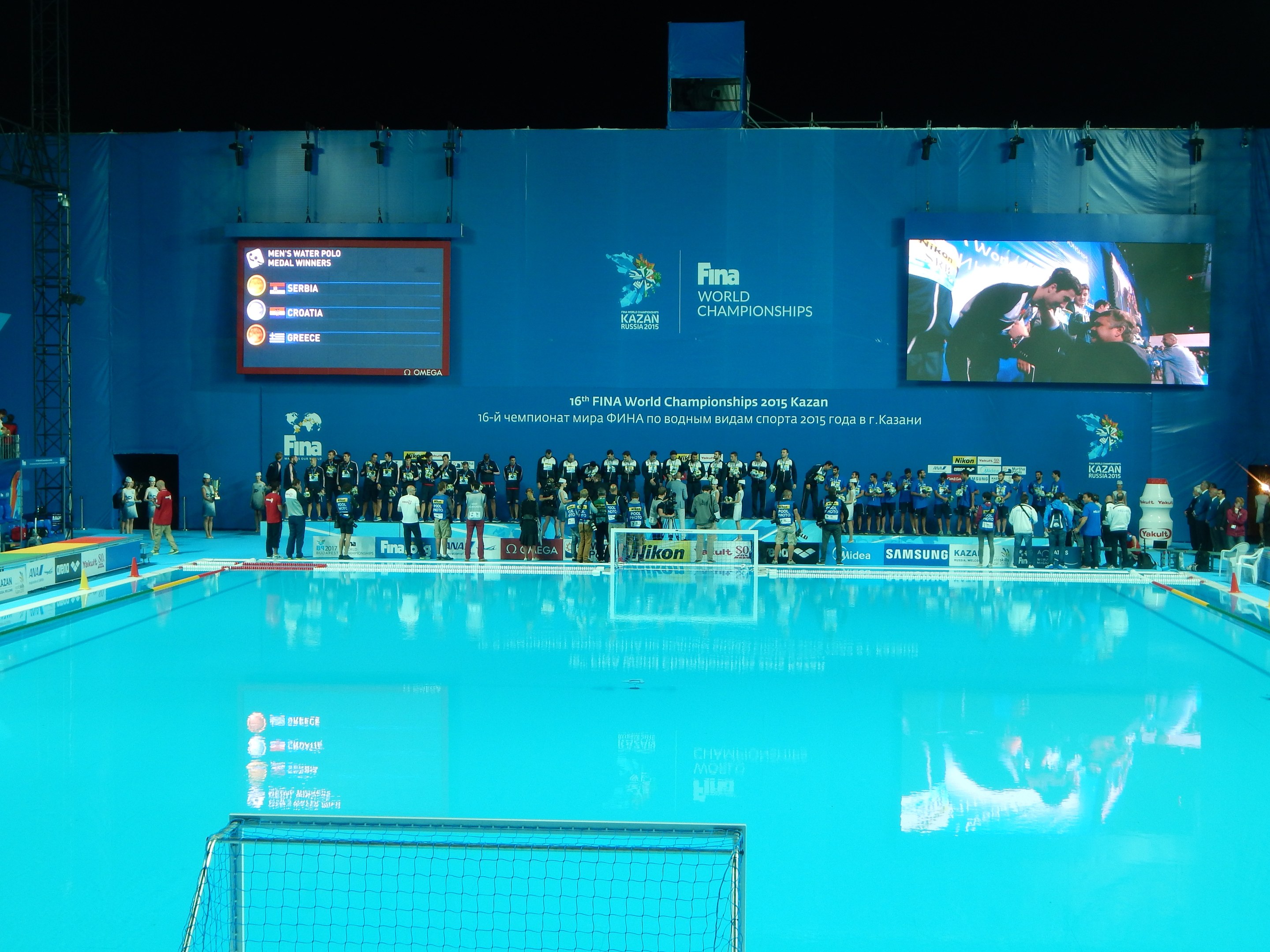 Gruppe von Menschen, die um ein Hallenbad mit Netz am Boden stehen, mit einer Wand im Hintergrund, die "FINA World Championships 2015 Kazan"-Text und Logos anzeigt, sowie Trägern und Lampen.