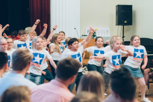Kinder in weißen Hemden und blauen Shorts tanzen auf einer Bühne vor sitzenden Zuschauern während eines internationalen Schul-Tanzwettbewerbs.