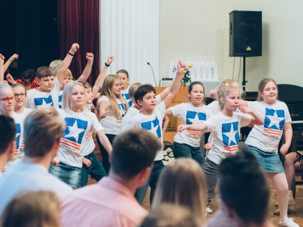 Kinder in weißen Hemden und blauen Shorts tanzen auf einer Bühne vor sitzenden Zuschauern während eines internationalen Schul-Tanzwettbewerbs.