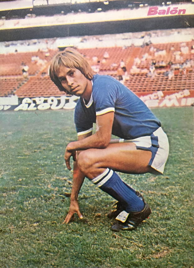 Ein Mann kniend auf einem Fußballfeld umgeben von Menschen auf Stühlen und Tafeln mit Text, tragend Schuhe.