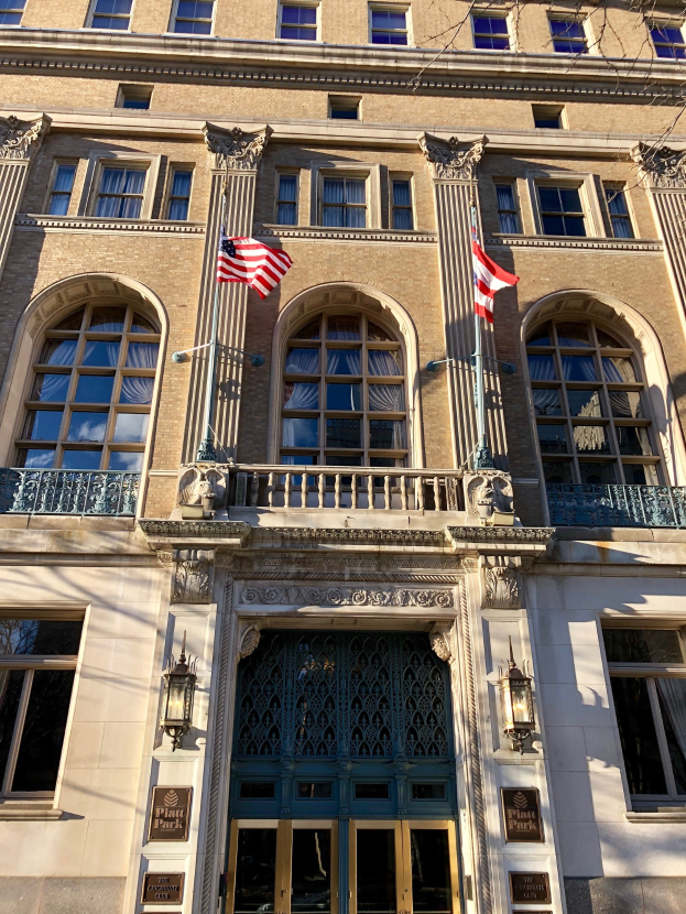 Außenansicht des Plaza Hotels in New York City, mit Glasfenstern, -türen, -säulen, -geländern, -leuchten, Texttafeln und Fahnen vor einem Himmelhintergrund.
