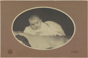 Ein altes Schwarz-Weiß-Foto eines Babys, das auf einem Bett liegt und von einer Decke bedeckt ist, mit Text und Logo unten.