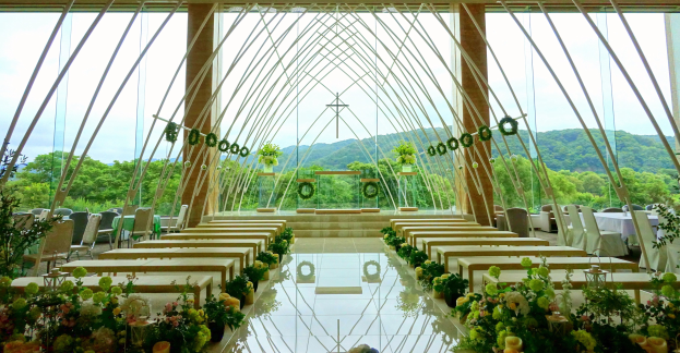 Eine Trauungszeremonie in einem großen Saal mit halbrunder Bestuhlung, geschmückt mit Blumenbouquets und anderen Gegenständen, vor einer Kulisse aus Bäumen, Hügeln und einem klaren blauen Himmel.