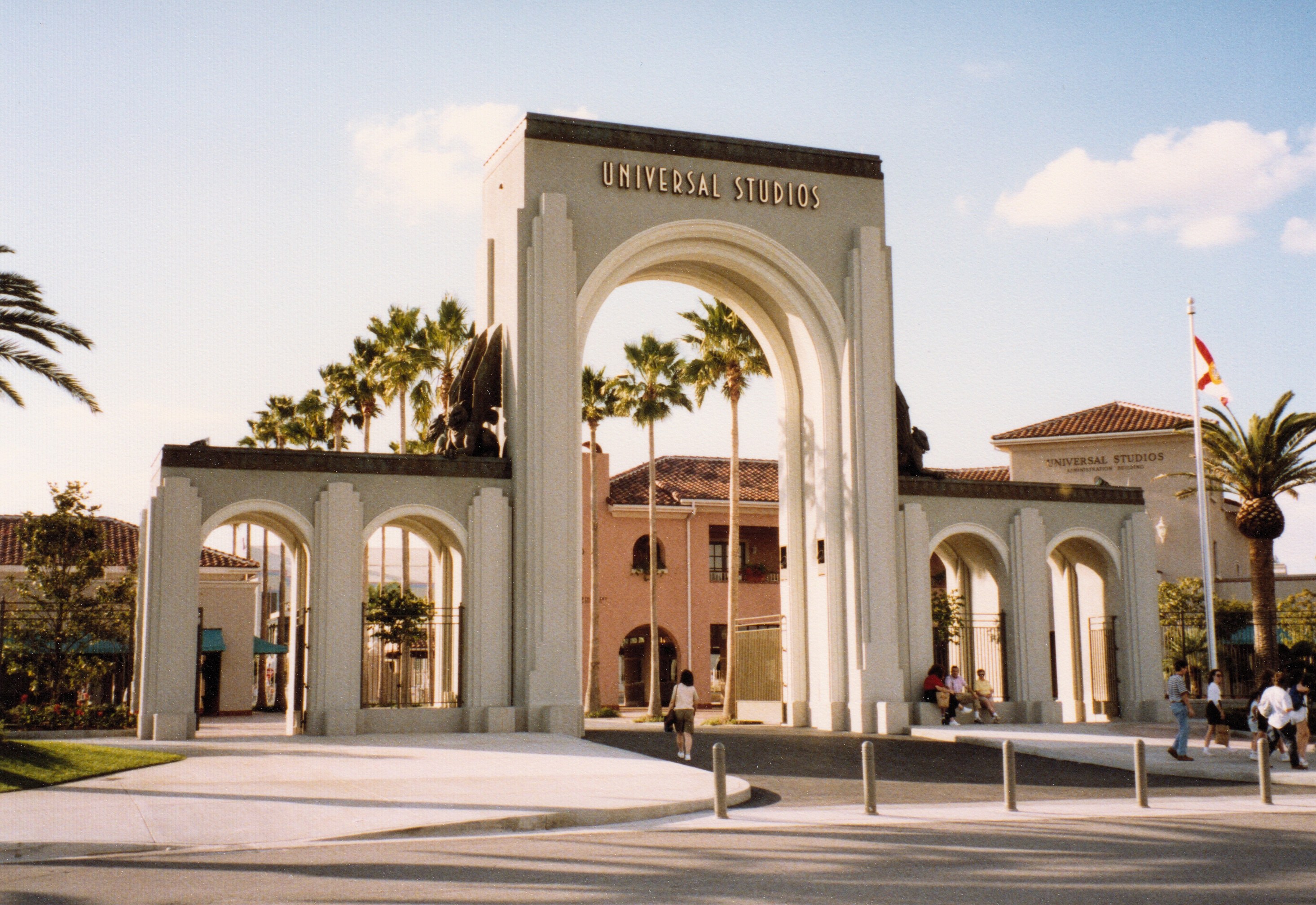 Eingang zu Universal Studios Hollywood mit einem Bogen, einer Statue, Menschen, Bäumen, Gebäuden, einem Fahnenmast und einem bewölkten Himmel.
