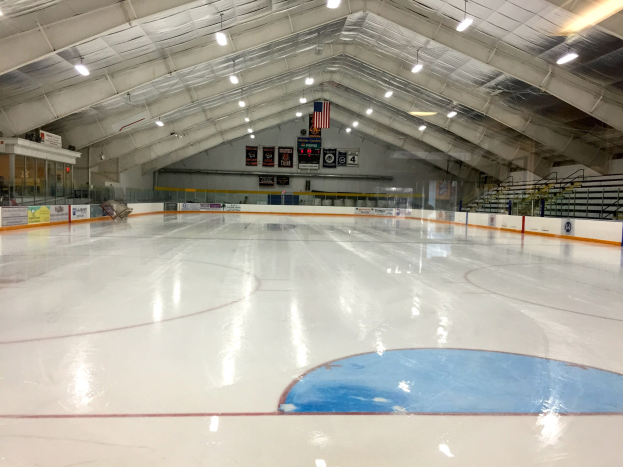 Große Indoor-Eisfläche mit Deckenbeleuchtung, Werbetafeln, einer Rückwand, Treppen mit Geländern und einigen Menschen, wahrscheinlich eine Hockey-Anlage.