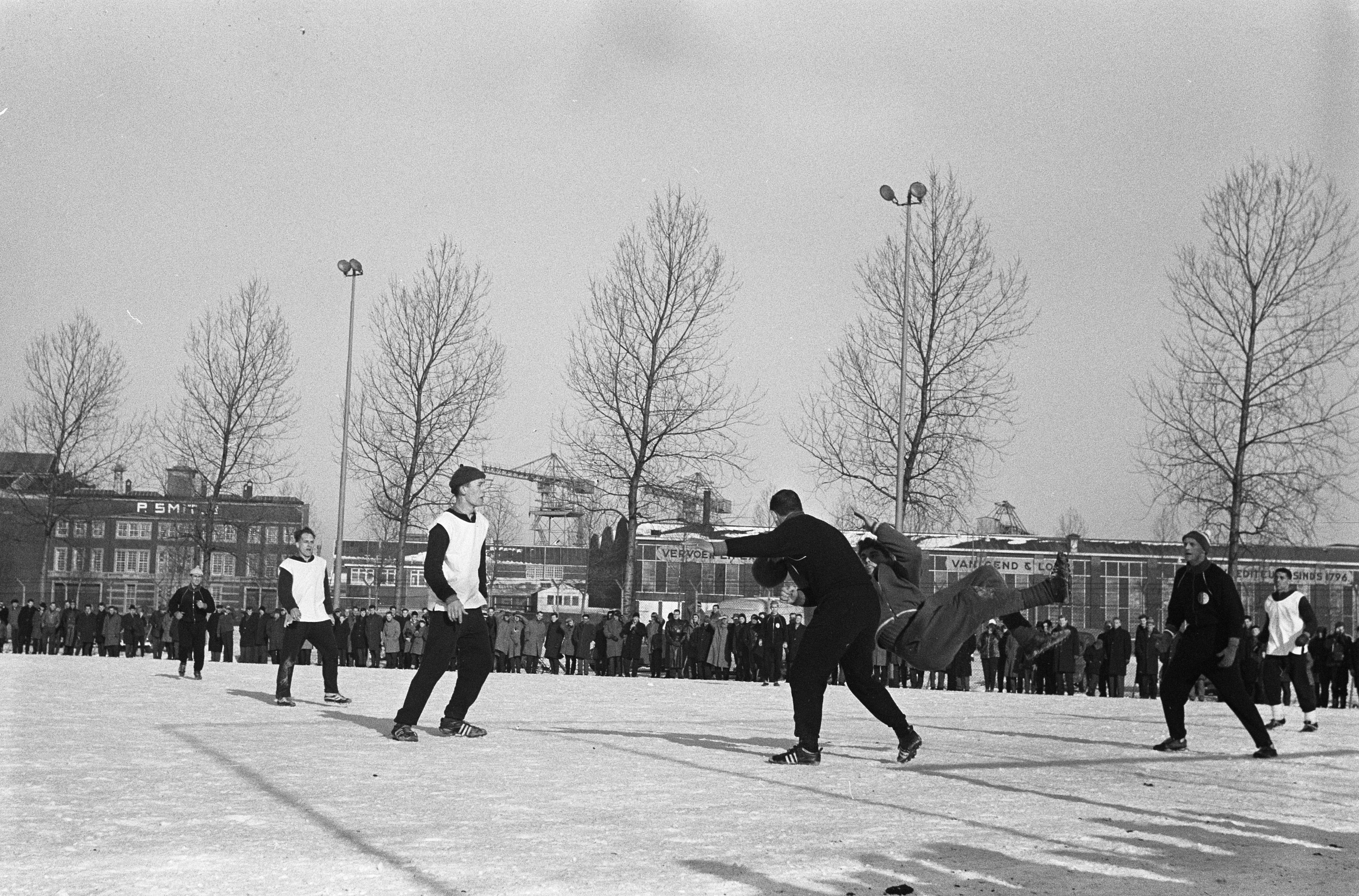 Eine Gruppe von Menschen spielt Eis-hockey im Schnee, mit Bäumen, Gebäuden, Laternenmasten und einem klaren Himmel im Hintergrund, dargestellt in Schwarz-Weiß.