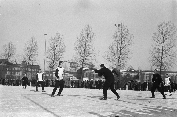 Eine Gruppe von Menschen spielt Eis-hockey im Schnee, mit Bäumen, Gebäuden, Laternenmasten und einem klaren Himmel im Hintergrund, dargestellt in Schwarz-Weiß.