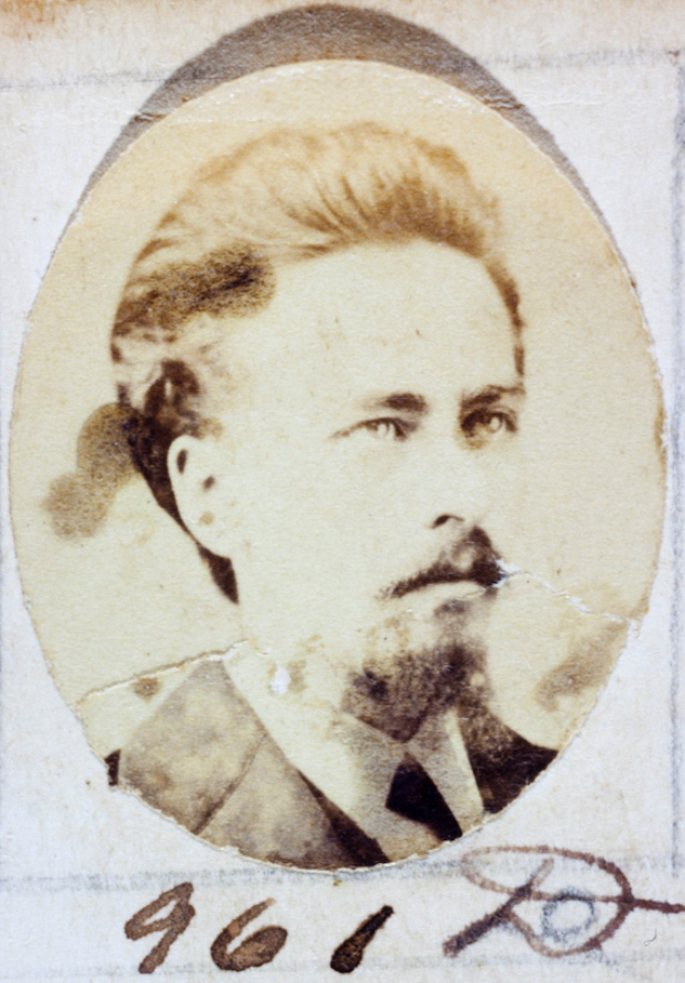 Ein altes Foto eines Mannes mit Schnurrbart und Bart, der einen Anzug und Krawatte trägt und direkt in die Kamera schaut.