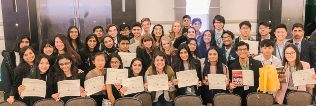 Gruppe von Menschen mit Urkunden bei einer Abschlussfeier, umgeben von Stühlen und einer Wand im Hintergrund.
