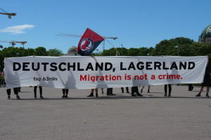 Eine Gruppe von Menschen, die draußen stehen und ein Banner halten, auf dem "Deutschland, Lagerland Migration ist kein Verbrechen" steht, sowie eine Flagge, mit Straßenlaternen, Bäumen, Kränen, einem Gebäude und einem bewölkten Himmel im Hintergrund.