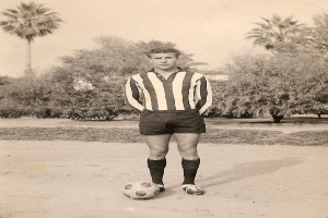 Ein Schwarz-Weiß-Foto eines älteren Mannes mit einem Fußball, vor einem Hintergrund aus Bäumen und Himmel.