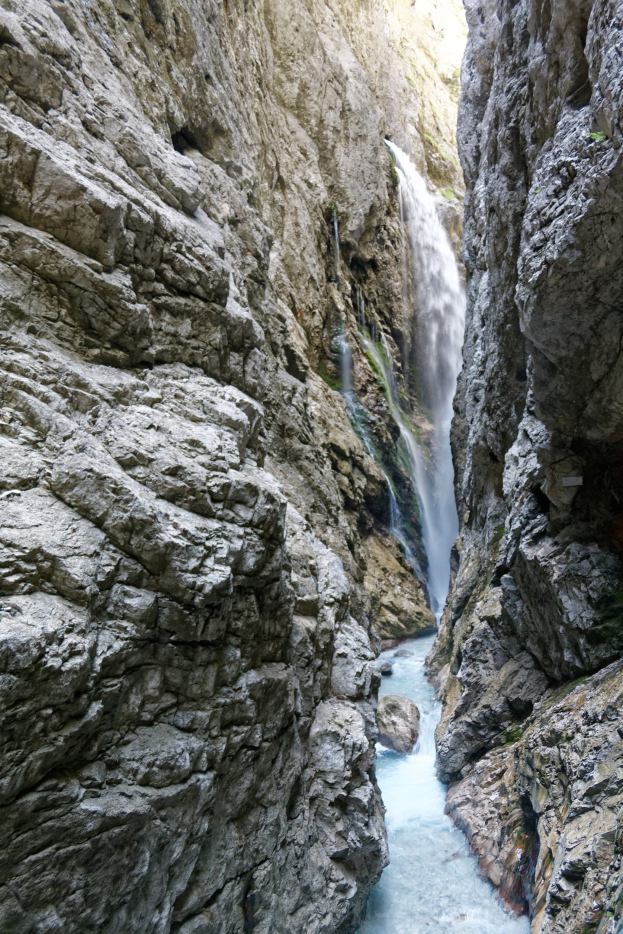 Ein kleiner Wasserfall stürzt eine steile, zerklüftete Schlucht mit schroffen, rauen Felsen hinab, umgeben von saftig grünen Hügeln unter strahlendem Sonnenschein.
