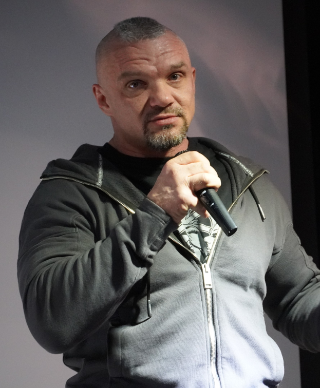 Ein Mann in einer schwarzen Kapuzenjacke und einem schwarzen Anzug mit einem weißen Hemd, glattem Haar und einem selbstbewussten Gesichtsausdruck, der ein Mikrofon hält, während er vor einer Wand steht.