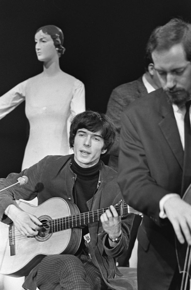 Ein Mann in einem schwarzen und weißen Outfit sitzt auf einem Stuhl und spielt eine Gitarre neben einer Schaufensterpuppe, mit einem Mikrofon davor, vor einem dunklen Hintergrund, als Hommage an den verstorbenen Paul McCartney.