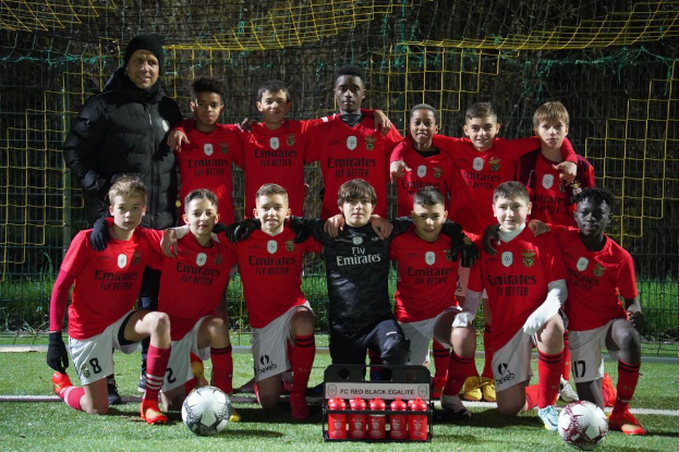 Eine Gruppe junger Jungs in roten T-Shirts und schwarzen Shorts posiert auf einem Fußballfeld für ein Foto, umgeben von mehreren Fußballen, einem Netz, einem Zaun und Bäumen im Hintergrund.