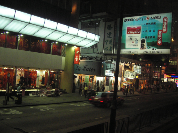Eine nächtliche Stadtstraße mit einem vorbeifahrenden Auto, erleuchteten Gebäuden, Schaufenstern und Fußgängern auf einem Gehweg unter einem dunklen Himmel.