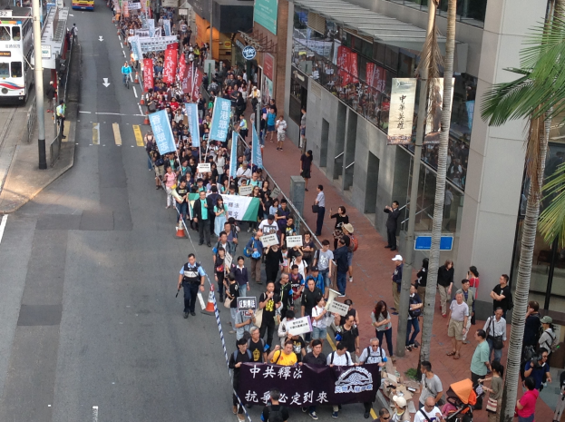 Großer Protestmarsch in Hong Kong mit Menschen mit Schildern und Plakaten, flankiert von Bäumen und Gebäuden auf der rechten Seite und Fahrzeugen auf der linken Seite.