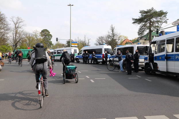 Eine Gruppe von Menschen, die auf Fahrrädern eine Straße entlangfahren, mit Polizeiwagen und anderen Fahrzeugen, einige tragen Helme und haben Taschen oder schieben Kinderwagen, mit Bäumen, Laternenmästen, Verkehrsampeln, Schildern, Gebäuden und einem bewölktem Himmel im Hintergrund.