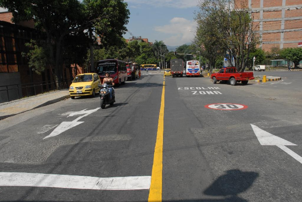 Eine Straßenszene mit Gebäuden, Bäumen, fahrenden Fahrzeugen, Verkehrsschildern, Ampeln, Warnmarkierungen und einem blauen Himmel.