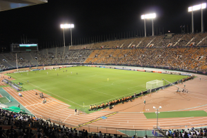 Ein großes, beleuchtetes Stadion voller Zuschauer bei einem nächtlichen Fußballspiel, mit einem Hintergrundscoreboard.