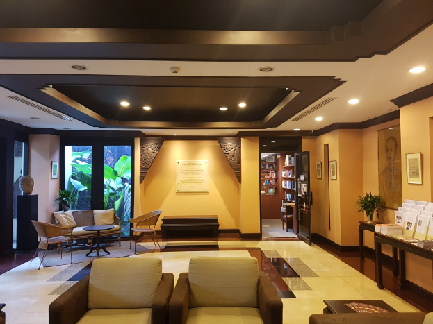 Ein geräumiger Hotelfoyer mit Sitzbereichen, Dekorationspflanzen, Bücherregalen, Wandkunst und Seitentisch-Dekoration, beleuchtet von elektrischem Licht und natürlichem Sonnenlicht durch Fenster und Türen.