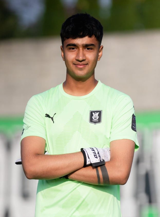 Ein junger Mann in einem grünen T-Shirt steht auf einem Fußballfeld mit verschränkten Armen und lächelt, wahrscheinlich ein Torwart, mit einem unscharfen Hintergrund.