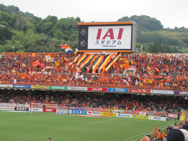 Fußballspiel in einem Stadion mit einer großen Zuschauermenge, saftigem Grün, Torstangen, Bannern, Fahnen, einem großen Bildschirm, Bäumen und einem klaren blauen Himmel.