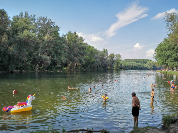 Menschen schwimmen in einem See umgeben von Grün, mit einer Person am Ufer und aufblasbaren Spielzeugen im Wasser unter einem klaren blauen Himmel.