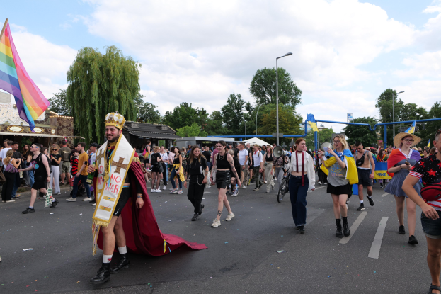 Eine Gruppe von Menschen marschiert bei der Pride Parade 2018 mit einer Regenbogenflagge und Musikinstrumenten, einige tragen Mützen, vor einem Hintergrund aus Laternenmasten, Bäumen, Schuppen und einem bewölkten Himmel.