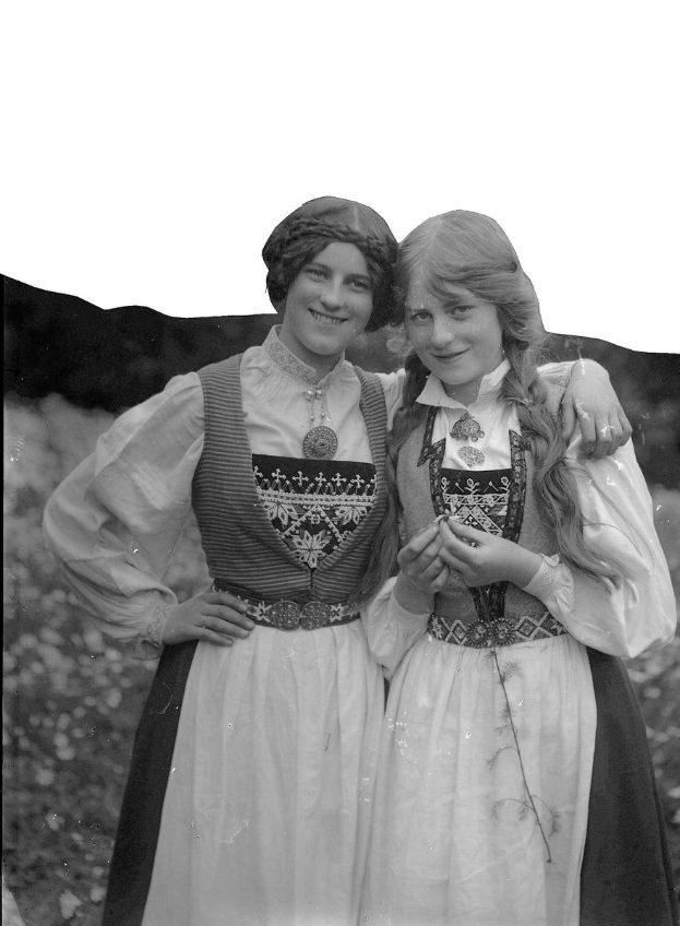 Zwei junge Frauen in traditionellen bayerischen Trachten mit weißen und schwarzen bestickten und perlenden Oberteilen stehen lachend vor einer Kulisse aus grünem Blattwerk und einem hellen Himmel.