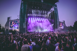 Eine große Menschenmenge bei einem Musikfestival, mit Performern auf einer zentralen Bühne, die von Scheinwerfern beleuchtet und von Bildschirmen umgeben ist, unter einem klaren blauen Himmel mit einem Gebäude und Bäumen im Hintergrund.