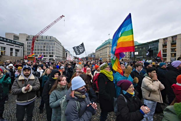 Eine große Gruppe von Menschen mit Fahnen und Transparenten vor einem Gebäude während einer LGBTQ+-Rechtsdemo in Berlin, mit Gebäuden, einem Kran und einem bewölkten Himmel im Hintergrund.
