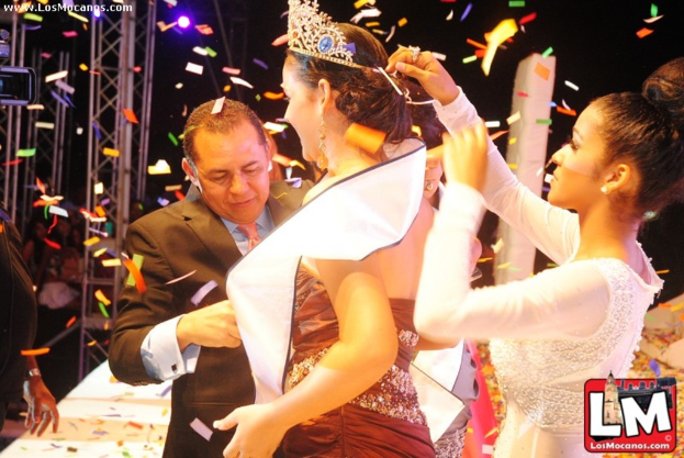 Ein Mädchen auf der Bühne bei einer Miss World Show mit einer Krone, dahinter ein Mädchen, das sie anpasst, und ein Mann in einem schwarzen Anzug, der vor ihr die Hand schüttelt, vor einem dunklen Hintergrund mit Metallrahmen.