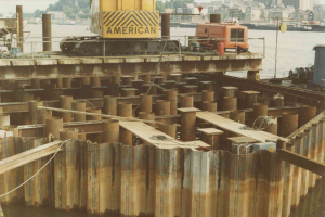 Baustelle mit einem Kran, der Stahlstangen aus einem Haufen hebt, umgeben von Wasser, Gebäuden, Bäumen und einem klaren blauen Himmel.
