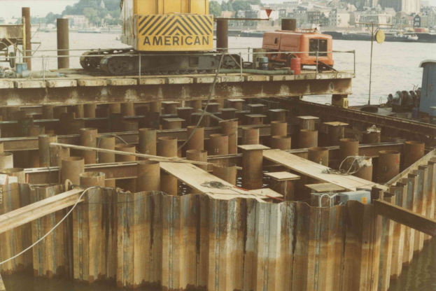 Baustelle mit einem Kran, der Stahlstangen aus einem Haufen hebt, umgeben von Wasser, Gebäuden, Bäumen und einem klaren blauen Himmel.