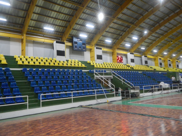 Großer Hallensportplatz mit Holzboden, blauen und gelben Sitzplätzen, Geländern, Treppen mit Geländern, wandmontierten Bildschirmen, Deckenleuchten und einigen anwesenden Personen.