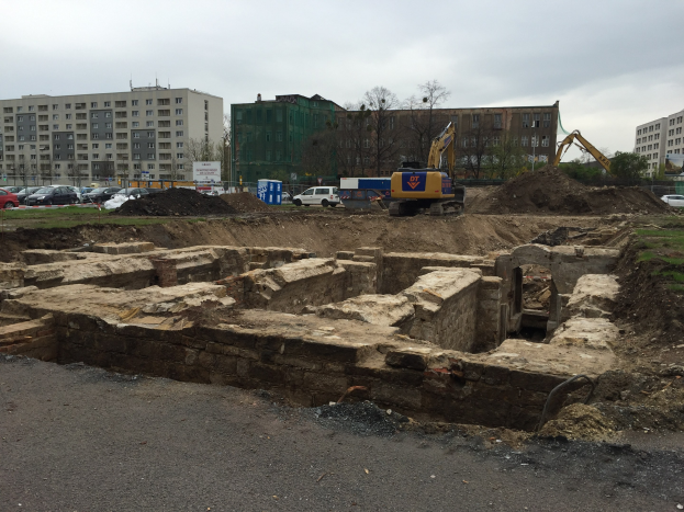 Baustelle mit einem großen Erdberg, Baggern, Fahrzeugen, Gras, Bäumen, Gebäuden mit Fenstern im Hintergrund, bewölktem Himmel und einer Straße unten.