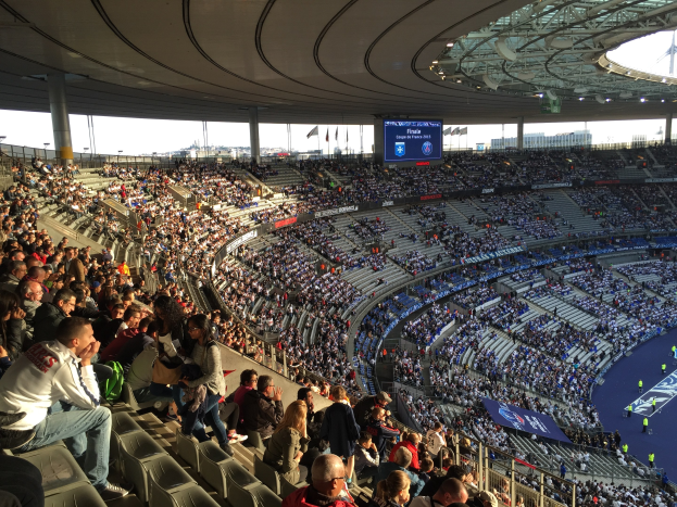 Große Menschenmenge in einem Stadion bei einem Fußballspiel mit einer Bühne, Flaggen, Stangen und einem Bildschirm im Hintergrund, identifiziert als Allianz Arena in München, Deutschland.