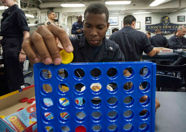 Ein Mann spielt Connect 4 an einem Tisch, umgeben von Menschen, mit Kisten und anderen Gegenständen auf dem Tisch und Menschen an Tischen im Hintergrund, Fotorahmen an der Wand und Lampen an der Decke.
