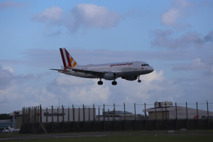 Germanwings Airbus A320-200 beim Abheben vom Frankfurt Airport, mit Gras darunter, einem Zaun im Vordergrund, Gebäuden und Bäumen im Hintergrund und einer bewölkten Himmel.